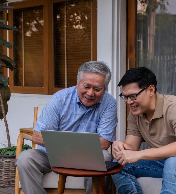 Happy Asian family with technology at home. Adult son teach senior father using laptop computer with internet for online shopping or social media at home terrace. Family enjoy and having fun weekend activity together with happiness